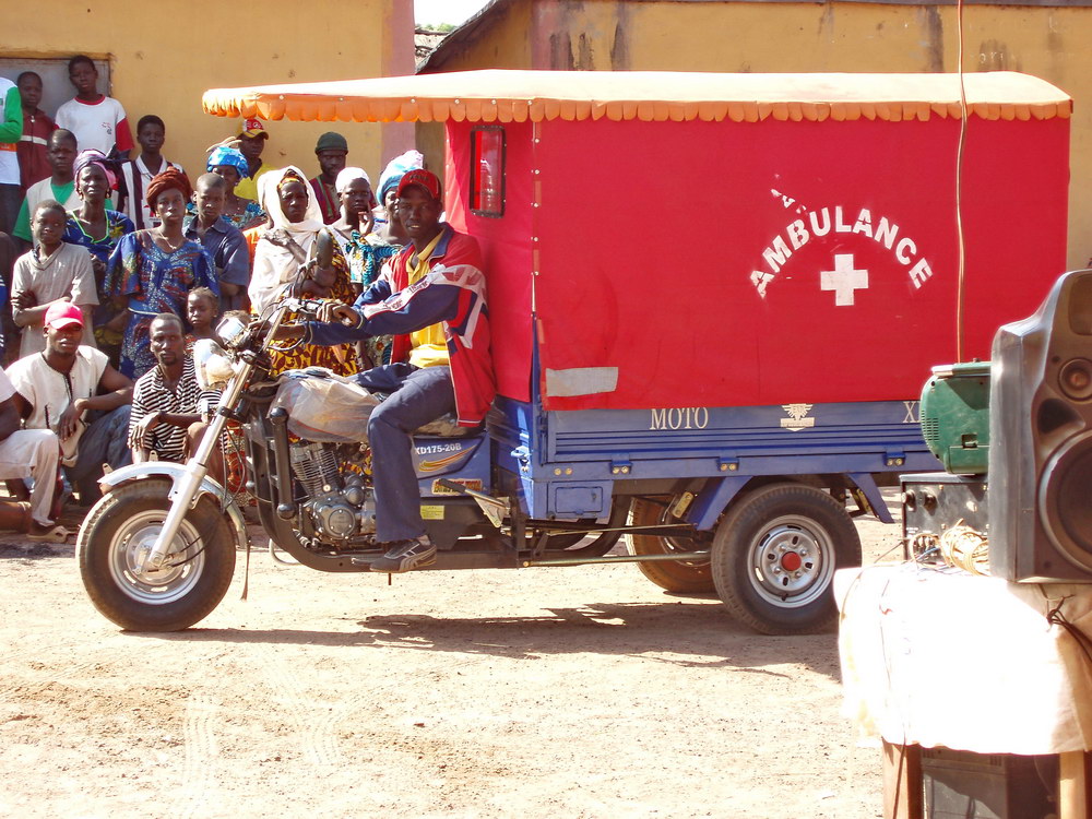 Red Ambulance trailer attached behind the back of a motorbike, watched by a crowd of village women