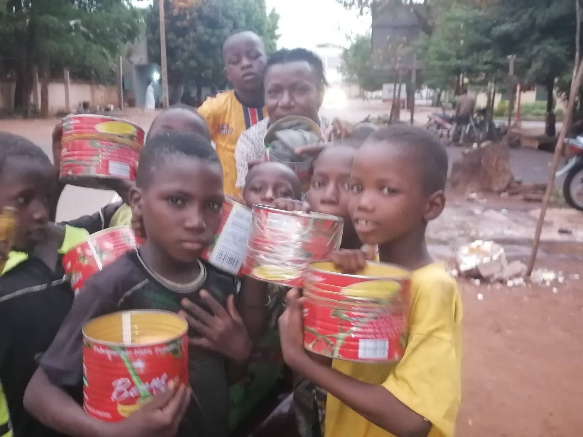 Group of boys with red tomato paste tins with man on unpaved, tree lined street