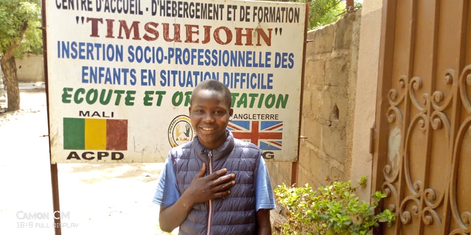 Young boy with hand on his heart standing in front of partner office sign board showing Mali and UK flags